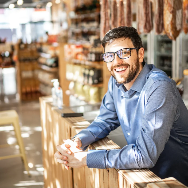 Managed-It-Services-Broward-County A smiling male business owner with glasses leaning on a wooden counter inside a specialty food market with cured meats hanging in the background.