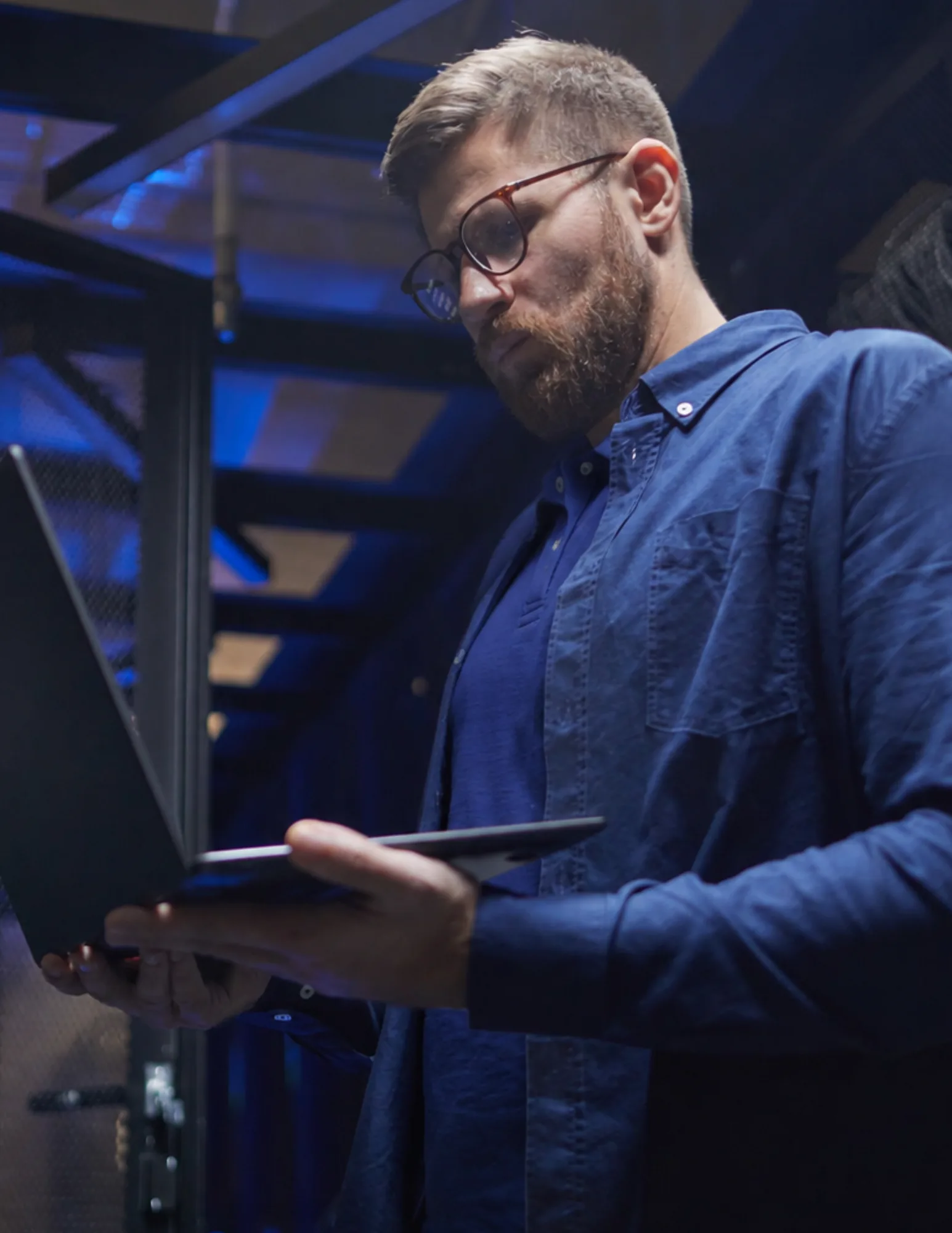 A bearded man in a light gray sweater typing at a desktop computer monitor in a bright office, with a woman working at a separate desk in the background.