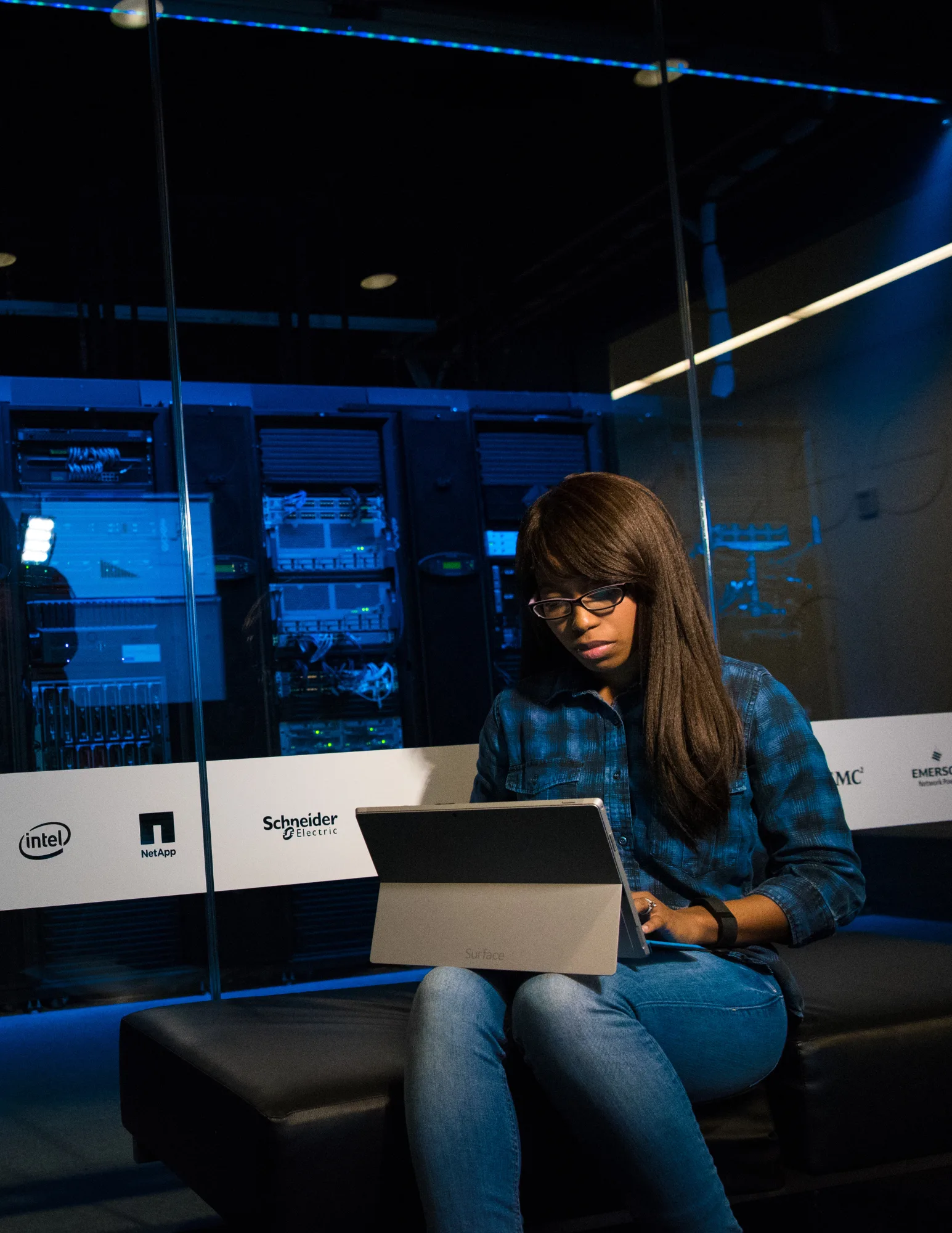 Business-Cybersecurity-Broward-County A woman wearing glasses and a blue plaid shirt sitting on a black bench in a server room, working on a Surface tablet with glowing blue equipment racks in the background.