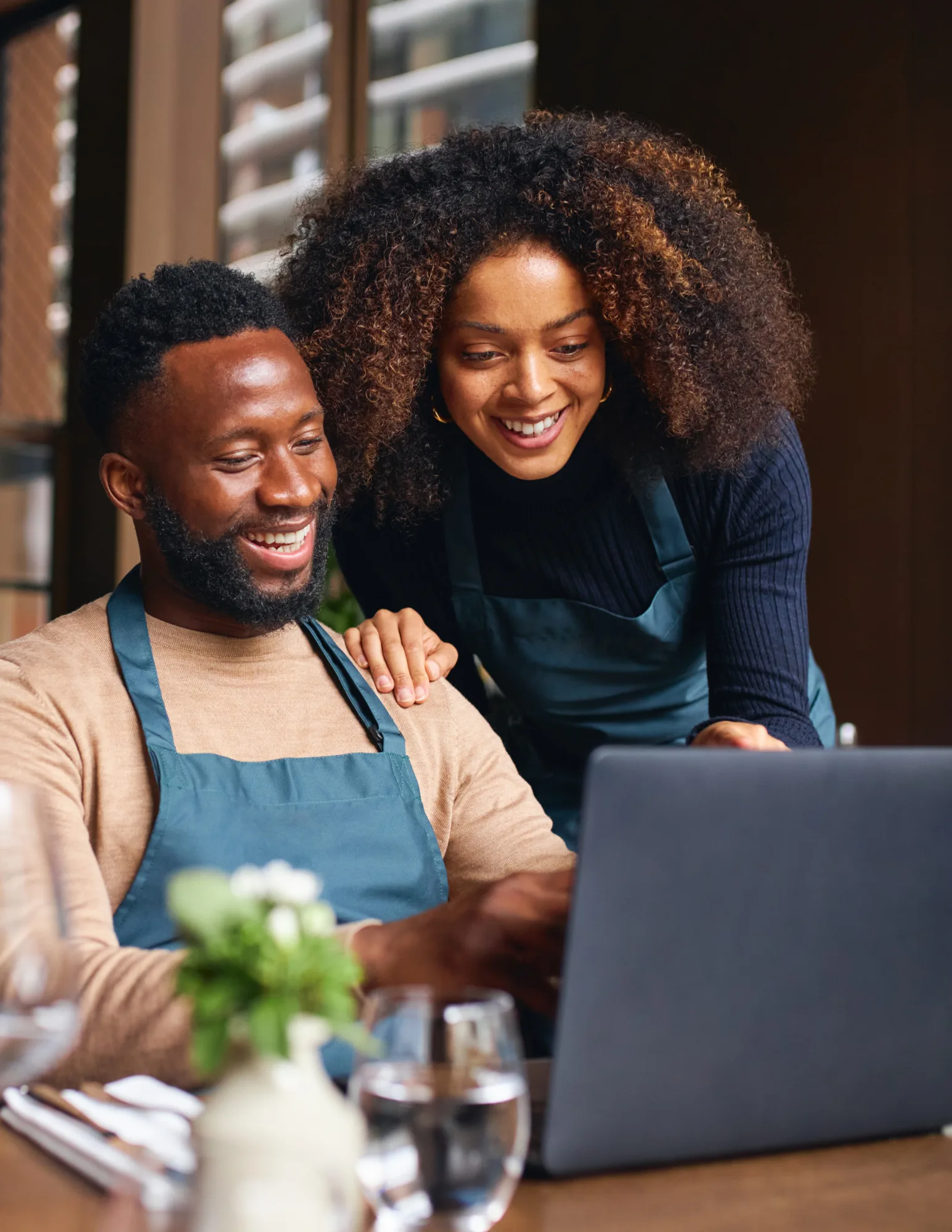 Local-It-Support-Team-Broward-County A male and female restaurant owner wearing green aprons smiling together while looking at a laptop on a set dining table.