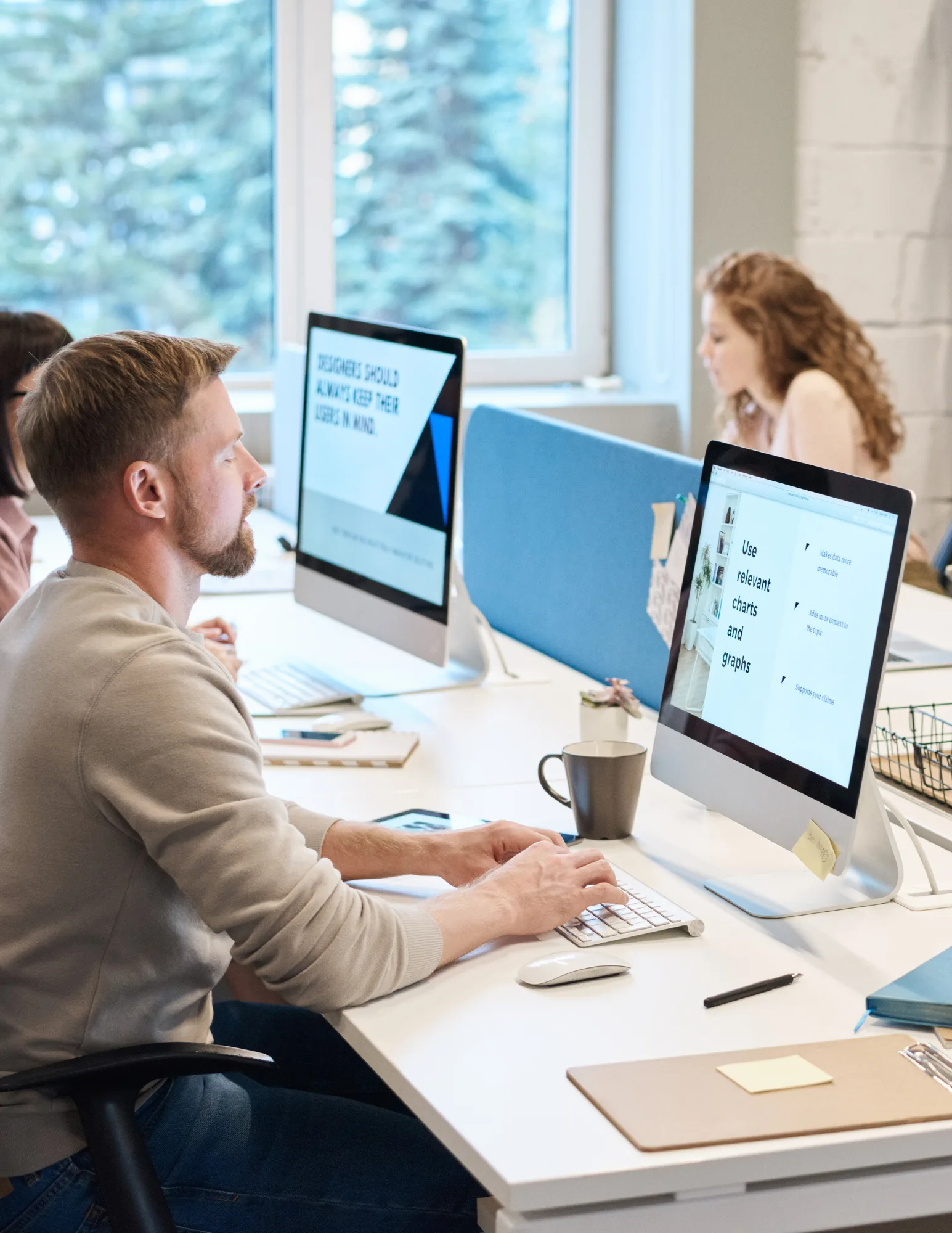 Proactive-It-Support-Broward-County A bearded man in a light gray sweater typing at a desktop computer monitor in a bright office, with a woman working at a separate desk in the background.